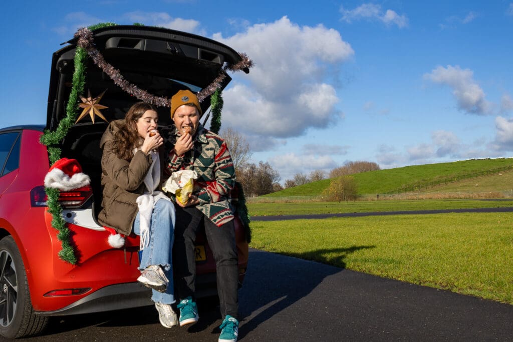 Twee mensen die in de achterbak van een auto oliebollen eten tijdens De Oliebollen Drive-in